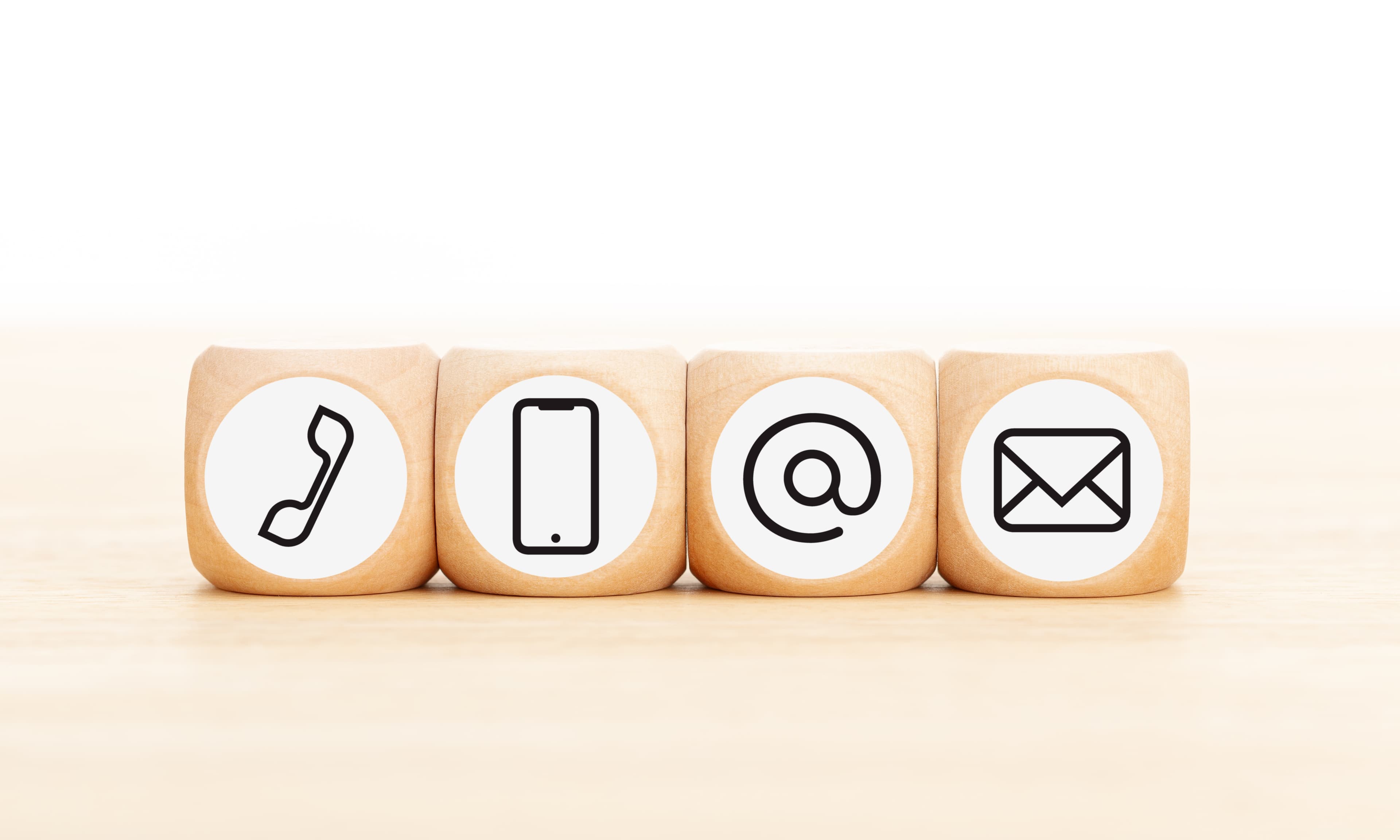 Vier Holzwürfel mit Symbolen für Telefon, Smartphone, At-Zeichen und E-Mail nebeneinander auf einer Holzoberfläche.